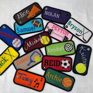 Könnte beinhalten: Eine Sammlung bunter personalisierter Schlüsselanhänger mit gestickten Namen und Sportmotiven. Die Schlüsselanhänger zeigen verschiedene Sportarten, darunter Baseball, American Football, Basketball, Volleyball, Hockey, Golf, Lacrosse, Gewichtheben und Musik.
