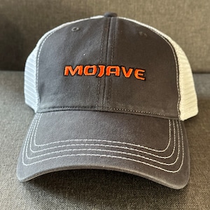 May include: A gray and white baseball cap with a black mesh back. The front of the cap has the word "MOJAVE" embroidered in orange.