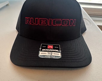 May include: Black trucker hat with a red embroidered "RUBICON" logo on the front. The hat has a black mesh back and a black adjustable strap with a red "RICHARDSON" label.