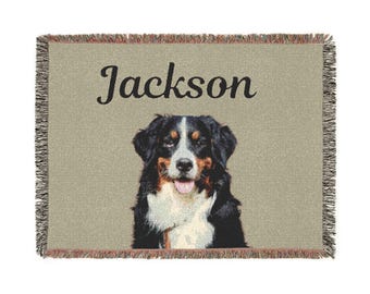 May include: A woven throw blanket featuring a Bernese Mountain Dog portrait with the name "Jackson" in black script. The dog has black, white, and brown markings. The blanket has a tan background and fringed edges.