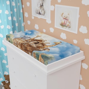 Könnte beinhalten: Eine weiße Wickelkommode mit einer farbenfrohen Wickelauflage mit einer Illustration der Arche Noah mit Giraffen, Elefanten und anderen Tieren. Der Hintergrund ist ein blauer Himmel mit Wolken. Die Wickelkommode befindet sich in einem Kinderzimmer.