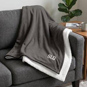 Könnte beinhalten: Eine dunkelgraue Decke mit weißem Sherpa-Futter liegt über einem grauen Sofa. Die Decke hat die gestickten Initialen "SLD" in Weiß. Ein gemütliches Wohnaccessoire.