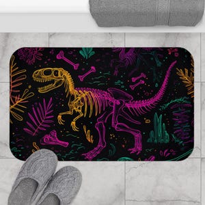 May include: Black rectangular bath mat featuring a neon dinosaur skeleton design. The dinosaur skeleton is outlined in yellow and pink, with green and orange accents. The background is black with colorful plant illustrations.