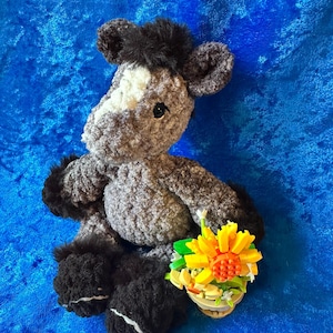 May include: A handmade crochet horse plush toy in shades of gray, black, and white, with a small flower arrangement. The horse has a white patch on its face and black mane and hooves. The flower arrangement features yellow and orange flowers.