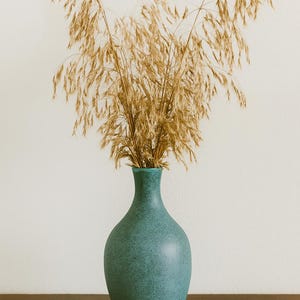 May include: A teal ceramic vase with a speckled texture holds dried ornamental grass. The vase has a rounded body and a narrow neck. The grass is a light brown color and adds a natural element to the composition.