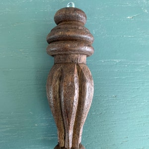 May include: A dark brown, carved wooden furniture leg with a decorative design. The leg features a rounded top, a series of stacked, ring-like sections, and a carved, fluted lower portion. A small, clear loop is attached to the top.