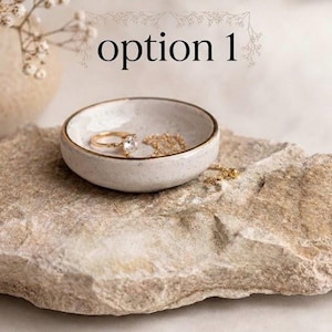 May include: A small, round, speckled off-white ceramic trinket dish with a thin gold-toned rim. Inside the dish sits a gold ring with a clear stone and a delicate gold chain. The text "option 1" is visible above the dish in a serif font.