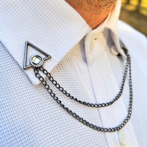 May include: A silver collar pin with a geometric triangle design and a clear rhinestone. The pin is attached to a double chain.