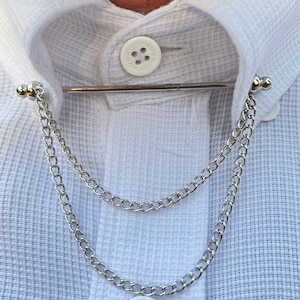 Op de afbeelding: Een zilveren kraagspeld met een dubbele ketting eraan. De kraagspeld zit op een wit overhemd.