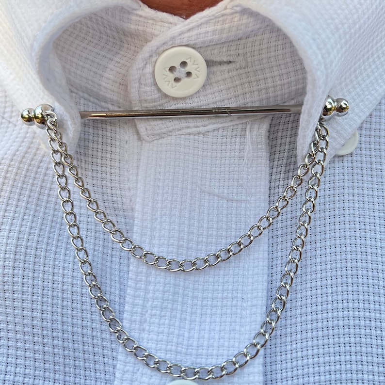 Op de afbeelding: Een zilveren kraagspeld met een dubbele ketting eraan. De kraagspeld zit op een wit overhemd.