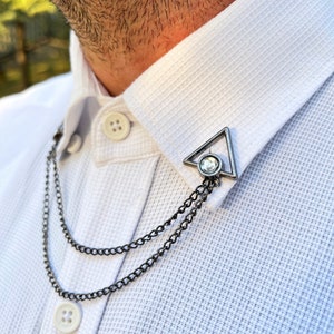 May include: A silver collar pin with a clear rhinestone in the center. The pin is in the shape of a triangle and is attached to a double chain.