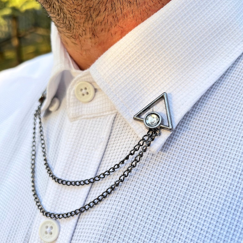 May include: A silver collar pin with a clear rhinestone in the center. The pin is in the shape of a triangle and is attached to a double chain.