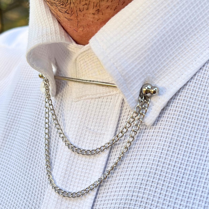 Op de afbeelding: Een zilveren kraagspeld met een ketting aan elk uiteinde. De ketting is dubbel en heeft aan elk uiteinde een ronde, zilveren kraal.