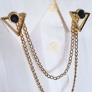 May include: A gold chain with two triangular pendants featuring black circular accents. The chain is draped over a white collared shirt.