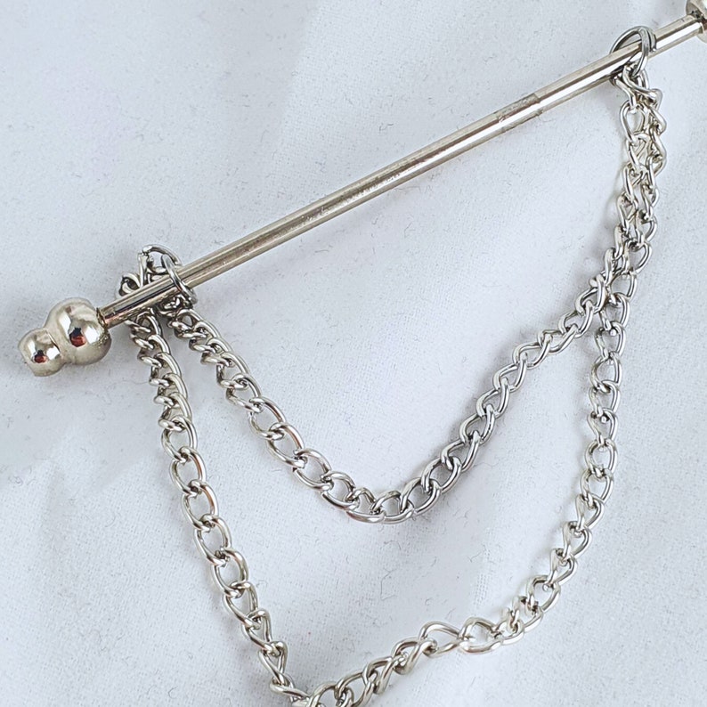 Op de afbeelding: Zilveren stropdasgesp met een eraan bevestigde ketting. De ketting heeft twee strengen en is met een kleine bal aan het uiteinde aan de gesp bevestigd.