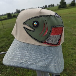 May include: A beige baseball cap featuring a decorative applique of a fish on the front panel. The fish is olive green with a red and silver textured body and a circular iridescent eye. The cap has a light blue denim brim with white stitching.