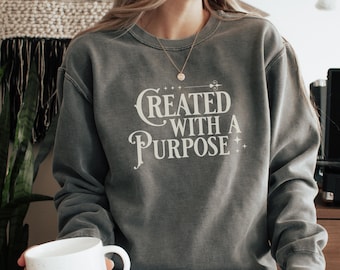 May include: A grey sweatshirt with the text "Created with a Purpose" in white lettering. The sweatshirt has a distressed look and is being worn by a person.