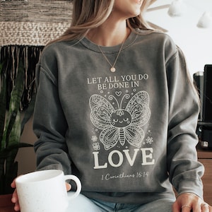 Puede incluir: Una sudadera gris antracita con un diseño de mariposa blanca y el texto "Let all you do be done in love 1 Corinthians 16:14".