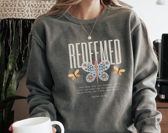 May include: A grey sweatshirt with the word "REDEEMED" in white block letters. A butterfly with blue, orange, and white details is centered below the word. The sweatshirt also features text from Isaiah 44:22.