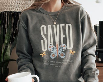 May include: A grey sweatshirt featuring the word "SAVED" in large white letters. A butterfly design with floral accents is centered below the word. The text "For God so loved the world..." and "JOHN 3:16" are printed below the butterfly.