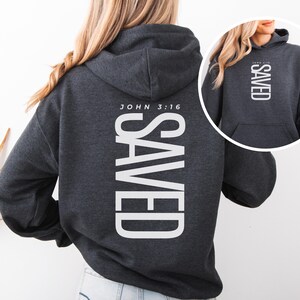 May include: Dark gray hoodie with the word "SAVED" printed vertically in large white letters on the back. The text "JOHN 3:16" is printed above the word "SAVED". A smaller image of the hoodie is in the upper right corner.