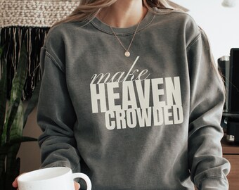 May include: A grey crew neck sweatshirt with the white text "make HEAVEN CROWDED". The sweatshirt is made of a soft material. A white mug is held in the lower left corner. The overall style is casual.