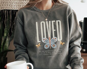May include: Grey sweatshirt with the word "LOVED" printed in white. A butterfly graphic with floral accents is below the word. The scripture "But God demonstrates his own love for us in this: While we were still sinners, Christ died for us. Romans 5:8" is printed below the butterfly.