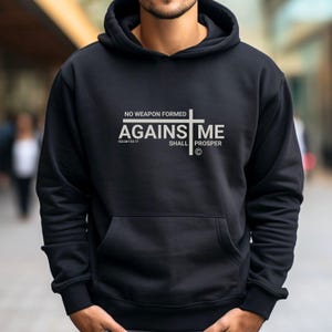 Może przedstawiać: Czarna bluza z kapturem z białym nadrukiem graficznym, na którym widnieje napis "No weapon formed against me shall prosper" wraz z krzyżem i wersetem z Księgi Izajasza 54:17.