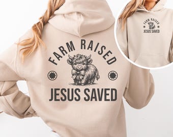 May include: A beige hoodie with a graphic of a Highland cow wearing a bow tie, with the text "FARM RAISED" above and "JESUS SAVED" below. A smaller image on the right shows the same design. The hoodie has a hood and a front pocket.