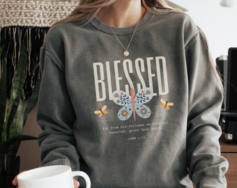 May include: A grey sweatshirt with the word "Blessed" printed in white, featuring a butterfly graphic and a scripture quote from John 1:16.