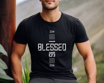 Puede incluir: Una camiseta negra con un diseño gráfico blanco de una cruz con la palabra "BLESSED" y los números "1:16" dentro de la cruz. El texto "JOHN" aparece arriba y abajo de la cruz.