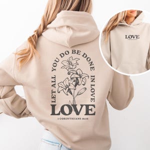 Puede incluir: Sudadera con capucha beige con un diseño floral y el texto "Que todo lo que hagas se haga con amor" y "LOVE" con la referencia bíblica "1 Corintios 16:14".