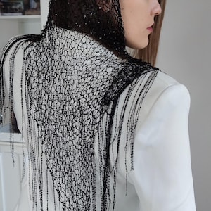 May include: A black, beaded head covering and shoulder wrap with fringe detail. The intricate design is draped over a white blazer. The head covering is worn by a person with long brown hair.