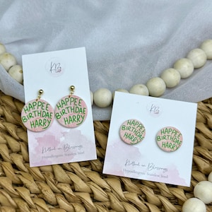 May include: Pink and green "Happee Birthdae Harry" earrings and studs. The earrings have a gold-coloured post. The text is in green on a pink background. The earrings and studs are on white cards.