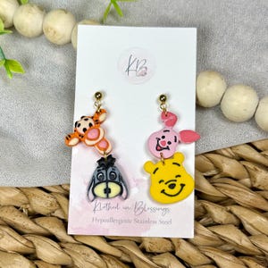 May include: Dangle earrings featuring cartoon characters. One earring has a Tigger, Eeyore, and a gold stud. The other earring has a Piglet, Winnie the Pooh, and a gold stud. The earrings are attached to a white card with text.