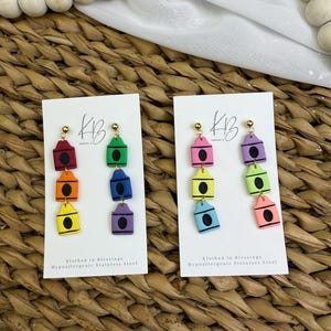 May include: Two pairs of colorful crayon-shaped earrings. Each earring features three stacked crayon shapes in different colors, including red, orange, yellow, green, blue, and purple. The earrings are on white cards with the text "Klothed in Blessings Hypoallergenic Stainless Steel".