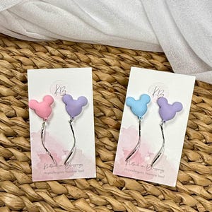 May include: Earrings with a Mickey Mouse head design in pink, lilac, and blue. Each earring has a curved silver-tone dangle. The earrings are displayed on white cards with the text "Clothed in Blessings" and "Hypoallergenic Stainless Steel".