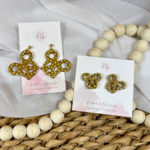 May include: Two pairs of gold-coloured pretzel-shaped earrings with white accents, displayed on white cards. The cards have a pink banner with the text "Klothed in Blessings" and "Hypoallergenic Stainless Steel".