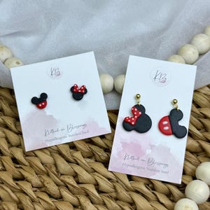 May include: Two pairs of black, red, and white polymer clay earrings. One pair is stud earrings with a Mickey Mouse design. The other pair is dangle earrings with a Mickey Mouse design. The earrings are displayed on white cards with text that reads "Clothed in Blessings" and "Hypoallergenic Stainless Steel."