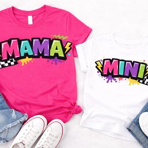 May include: Two t-shirts, one pink and one white, with the words "MAMA" and "MINI" in colorful, bold lettering. The shirts have a retro design with lightning bolts and paint splatters. The shirts are paired with denim shorts and white sneakers.