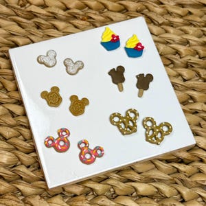 May include: A selection of miniature food-themed earrings, including cupcakes, ice cream bars, pretzels, and Mickey Mouse-shaped cookies. The earrings are displayed on a white surface, with a woven background.