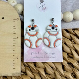 May include: A pair of white and orange BB-8 droid earrings. The earrings are on a white card with the text "Nothed in Blessings" and "Hypoallergenic Stainless Steel". The earrings are hanging from silver hooks.
