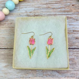 May include: A pair of gold-toned earrings with pink and green enamel tulip designs. The earrings are displayed in a white box with a light brown background.