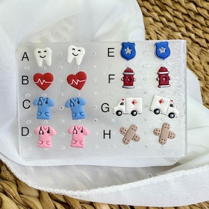 May include: A collection of medical-themed stud earrings. The earrings include designs of smiling teeth, hearts with a heartbeat, blue and pink scrubs, police badges, red fire hydrants, ambulances, and bandages. The earrings are displayed on a clear plastic card.