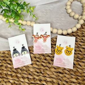 May include: Three pairs of character earrings on display. One pair features Eeyore, another Tigger, and the last pair features Winnie the Pooh. Each pair is on a pink and white card with the text "Klathed in Blessings" and "Hypoallergenic Stainless Steel".