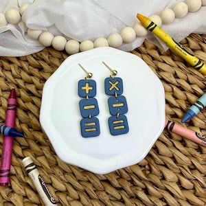 Puede incluir: Un par de pendientes azules con símbolos matemáticos dorados, incluyendo signos de suma, resta e igual, se exhiben en un plato blanco. Los pendientes son colgantes y tienen un poste dorado. Los crayones de varios colores rodean el plato.