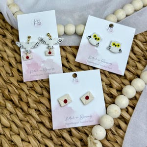 May include: Earrings with various designs, including white owls with yellow eyes and black markings, white owls with wings, and cream-colored envelopes with red seals. The earrings are displayed on white cards.