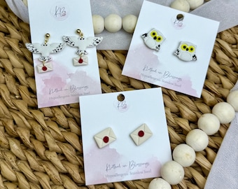 May include: A selection of earrings featuring owl and envelope designs. The owls are white with black markings and yellow eyes. The envelopes are cream-colored with red seals. The earrings are displayed on white cards.