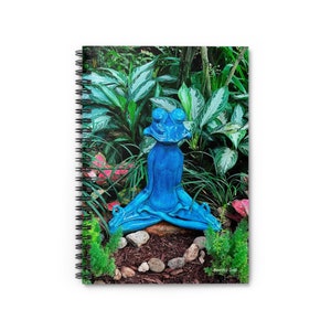 Peut inclure: Une sculpture de grenouille en céramique bleue en position de yoga, entourée de plantes vertes et de paillis brun. La grenouille est assise sur une petite bûche avec des pierres autour.