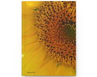 May include: A rectangular notebook featuring a close-up photograph of a vibrant yellow sunflower. The cover displays the text Annette's Soul in small black script in the bottom left corner, showcasing the intricate dark brown seed center.